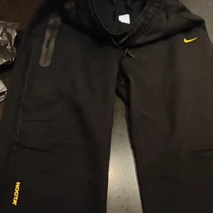 Svarta tech fleece byxor - Svarta mjukisbyxor med gul logga på benet och gul Swoosh vid höften. Byxorna har snörning i midjan och dragkedjeficka på sidan. Perfekta för en chill och sportig look.