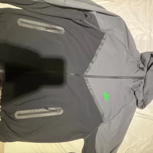 Nike svart och grå vindjacka med huva - Nike vindjacka i svart och grått med huva och hel dragkedja. Jackan har två vattentäta fickor med dragkedja och en grön Nike-logga på bröstet. Tillverkad i polyester, perfekt för blåsiga dagar. Sportig och clean look.