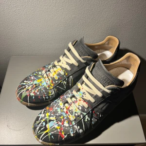 Maison Margiela sneakers med färgstänk - Unika svarta sneakers från Maison Margiela i skinn med coola färgstänk i gult, blått, rött och vitt. Beige snörning och klassisk gummisula med präglad logga undertill. Snygg streetstil med konstnärlig vibe, perfekt för dig som vill sticka ut.Man får box till och allt annat som hör till 