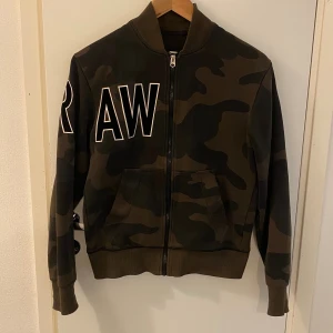 Camo bomberjacka från G-Star RAW XS - Snygg bomberjacka från G-Star RAW i camomönster med stora vita bokstäver på bröst och ärm. Jackan har dragkedja framtill, ribbade muddar och två fickor. Färgerna går i mörkgrönt och brunt, perfekt för en streetwear-look.