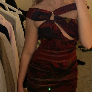 Vinröd kort klänning med cut out - Supersnygg vinröd kort klänning med unikt cut out-detalj framtill och twistad axelband. Klänningen har ett abstrakt mönster i mörka toner och sitter tight på kroppen. 