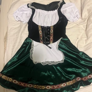 Grön och svart oktoberfest klänning  - Säljer en grön och svart oktoberfest klänning med kort puffärm, vit spetskant och snörning framtill. Kjolen har glansigt tyg och dekorativt band nertill samt ett vitt förkläde med spets. Perfekt för temafest eller maskerad!