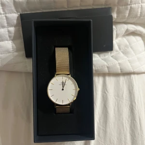 Daniel Wellington guldig meshklocka - Stilren klocka från Daniel Wellington med vit urtavla och guldig meshrem. Klockan har minimalistisk design med tunna guldiga markeringar och rund boett. Perfekt accessoar för en clean och modern look. I bra form!☺️