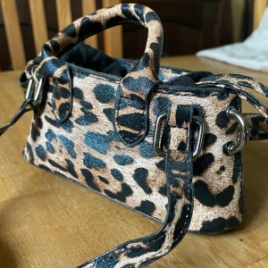 Leopardmönstrad axelväska med snörning - Axelväska med leopardmönster i beige, svart och brunt. Väskan har både handtag och axelrem, samt snygga metalldetaljer och snörning upptill. Perfekt storlek för det viktigaste och ger en edgy vibe till din outfit. 😍🤎