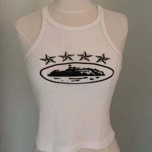 Vit Linnetopp med svart tryck - Vit ribbad linnetopp med svart tryck framtill som visar fyra stjärnor och ett motiv i en oval. Toppen har smala axelband och är croppad, perfekt för en trendig look. Materialet är stretchigt och bekvämt.
