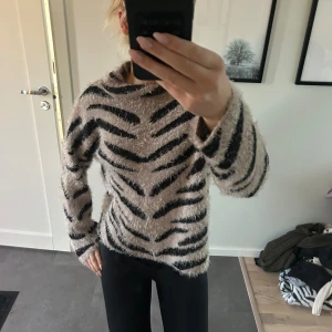 Zebramönstrad fluffig tröja - Supermjuk och fluffig stickad tröja från NA-KD i beige med svarta zebraränder. Tröjan har en lite högre hals och lång ärm. Bra skick och väldigt mysigt material.