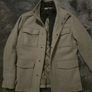 Beige rock från Jack & Jones XS - Snygg beige rock från Jack & Jones Premium i storlek XS. Jackan har fyra stora fickor framtill, hög krage och stängs med både dragkedja och knappar. Yttermaterialet är ullblandning och fodret är quiltat för extra värme. Perfekt för kyliga dagar.