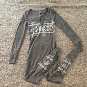 Grå onepiece från PINK Victoria’s secret  - Supermysig grå långärmad onepiece från PINK Victoria Secret med vintermönster och stor logga över bröstet. Ribbad bomullsmix, knappar framtill och dekorativa snöflingor på benen. Perfekt för kalla dagar och chill hemma. Aldrig använd.