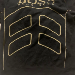 Svart hoodie från Hugo Boss med guldtryck - Svart hoodie från Hugo Boss med guldiga detaljer och tryck framtill. Tröjan har huva, dragkedja och långa ärmar. Snygg kontrast mellan svart och guld, perfekt för dig som vill sticka ut lite extra. Boss-logga syns tydligt på bröstet.