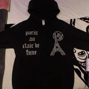 Svart hoodie med strass och Paris-tryck - Svart hoodie från BLINGLUCID i bomull och polyester med dragkedja, huva och glittrande strassdetaljer. Texten 'paris au clair de lune' och motiv av Eiffeltornet och måne i strass framtill. Perfekt statement-plagg för dig som gillar Paris-vibes.