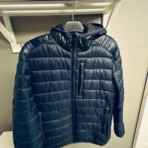 Mörkblå pufferjacka från Champion S - Säljer en mörkblå använd en gång på jag köpte en annan jacka pufferjacka från Champion i storlek S. Jackan har huva, dragkedja framtill och en bröstficka med dragkedja. Champion-loggan syns tydligt på ärmen och insidan. Perfekt för kalla dagar och riktigt snygg streetstil.