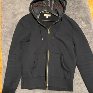 Svart hoodie från Burberry med dragkedja - Svart hoodie från Burberry med klassisk rutmönstrad huva, guldfärgad dragkedja och fickor framtill. Tröjan har långa ärmar, ribbade muddar och snörning i huvan. Perfekt för en chill och stilren look.