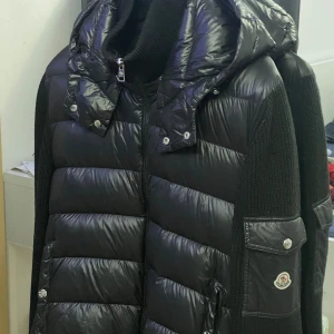 Svart Moncler pufferjacka med huva - Svart pufferjacka från Moncler med stickade ärmar och vadderad kropp. Jackan har en stor huva, dragkedja framtill och Moncler-logga på ärmen. Snygga detaljer med knappar och fickor på sidorna samt ärmen. Perfekt för kyliga dagar och riktigt stilren look. Storlek M passar S med kom med pris förslag 