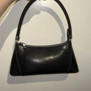 Svart axelväska från NA-KD - Snygg svart axelväska från NA-KD i stilren design med silverfärgade detaljer och dragkedja. Väskan är tillverkad i skinnimitation och har en praktisk ficka med dragkedja på baksidan. Perfekt storlek för det viktigaste och passar till många olika outfits.