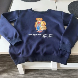 Blå Polo Bear tröja från Ralph Lauren - Säljer en mörkblå crewneck från Polo Ralph Lauren med ikoniska Polo Bear-trycket framtill. Tröjan har rund hals, ribbade muddar och är i mjuk bomullsmix. Perfekt för dig som gillar streetstyle och vill sticka ut med en klassisk men lekfull look. Vid några funderingar hör av dig// William 