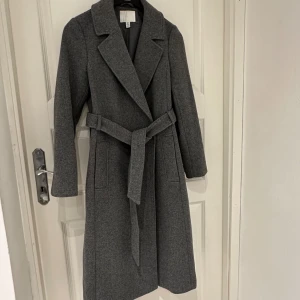 Grå kappa med bälte i ullblandning - Stilren grå kappa från H&M i ullblandning, med klassisk krage, knytskärp i midjan och två stora fickor framtill. Perfekt för kyliga dagar och ger en clean look. Fodrad insida för extra komfort.