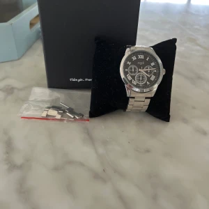 Regal silverfärgad klocka med svart urtavla - Stilren klocka från Regal med silverfärgad länk i metall och svart urtavla med romerska siffror. Klockan har flera små urtavlor och klassisk rund form. Perfekt accessoar för dig som gillar en tidlös look. Extra länkar medföljer.