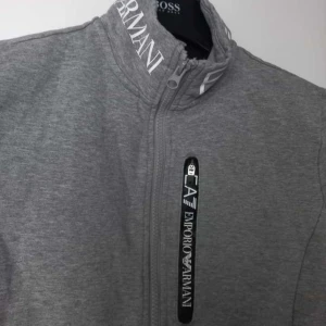 Grå zipjacka från Emporio Armani - Snygg grå jacka från Emporio Armani EA7 med hög krage och vit logga på kragen. Jackan har hel dragkedja framtill, lång ärm och en vertikal bröstficka med svart/vit EA7-detalj. Tillverkad i mjuk bomullsblandning, perfekt för en clean och sportig look.