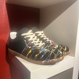 Svarta maison margiela sneakers  - Unika svarta sneakers med coola färgstänk i gult, rött, blått och vitt över hela ovandelen. Skorna har vit snörning och en klassisk gummisula i brunt. Materialet är skinn och designen är riktigt edgy med en konstnärlig vibe. Perfekt för dig som vill sticka ut med din stil. Ingen låda ingår, inga returer 
