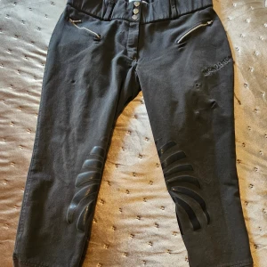 Svarta croppade byxor från Back2Back - Grå ridbyxor byxor från Back2Back med antiglid på benen. dragkedjor vid fickorna. Byxorna har hög midja med tre silverknappar och är gjorda i en stretchig mix av polyester, viskos och elastan. 