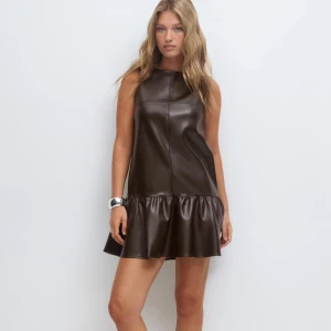 Brun kort klänning med volang - Snygg brun klänning i skinnimitation med volang nedtill. Modellen är ärmlös och har en rak passform som ger en cool och modern vibe. Perfekt för dig som gillar att sticka ut med stilrena detaljer och edgy material.Helt ny aldrig använd och är i storlek 36 säljer den för att den var för stor! 