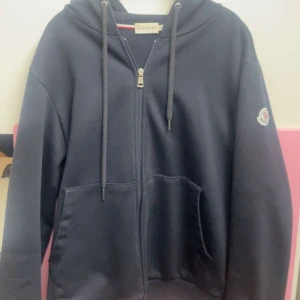 Svart hoodie från Moncler med dragkedja - Svart hoodie från Moncler med huva, snörning och dragkedja framtill. Tröjan har två stora fickor och Moncler-logga på ärmen. Perfekt för chill dagar och snygg streetstyle. Materialet är mjuk bomull och passformen är loose.
