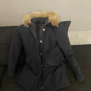Marinblå parkajacka Woolrich med äkta päls - Säljer en marinblå parkajacka med mysig äktapäls runt huvan. Jackan har långa ärmar, stora fickor framtill och stängs med både dragkedja och knappar. Perfekt för kalla dagar när du vill hålla dig varm och snygg.