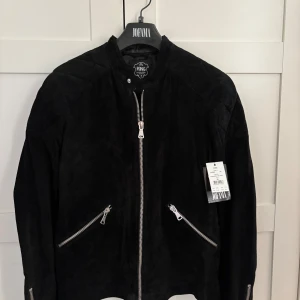 Svart mockajacka från Jofama x King - Svart mockajacka från Jofama x King med coola silvriga dragkedjor framtill och på ärmarna. Jackan har quiltade detaljer på axlar och ärmar samt en stilren ståkrage. Perfekt för dig som gillar en clean och edgy look. Ord pris 3495kr.