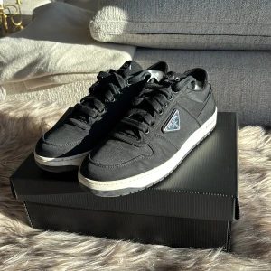 Svarta sneakers från Prada Milano - Snygga svarta sneakers från Prada Milano med klassisk låg siluett och vit sula. Skorna har snörning, rund tå och ikonisk Prada-logga i metall på sidan.  Pris 7500