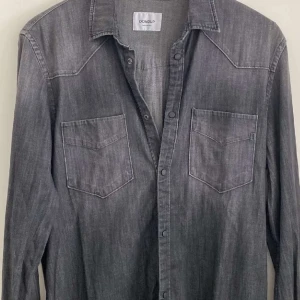 Dondup jeansskjorta  - Snygg grå jeansskjorta från Dondup med klassisk krage, två bröstfickor med lock och tryckknappar. Skjortan har lång ärm och coola sömdetaljer på axlarna. Perfekt för dig som gillar en avslappnad men stilren look. Skjortan är sparsamt använd, storlek M. Nypris ca 3K.
