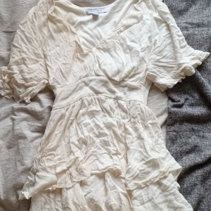 Krämvit kortklänning från Amisu - Supersöt krämvit kortklänning från Amisu med v-ringning, volangdetaljer och korta ärmar. Klänningen har en smickrande omlott-look upptill och är gjord i ett lätt, skrynkligt tyg som ger en avslappnad vibe. Perfekt för varma dagar och somriga tillfällen.