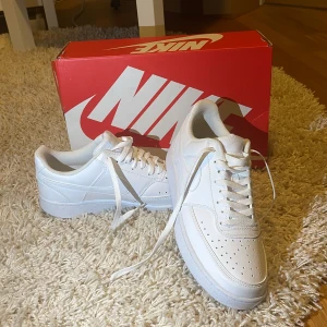 Nike Court Vision Low vita sneakers - Nike Court Vision Low sneakers i helvitt skinn med klassisk design och perforerade detaljer på tån. Snygga och stilrena med låg profil, snörning och rund tå. Perfekta för dig som gillar en clean look och vill ha ett par ikoniska Nike-sneakers.