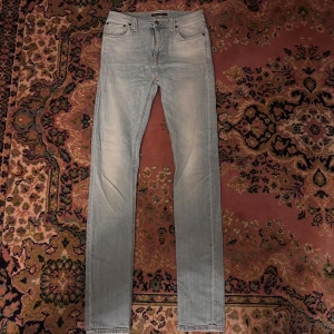 Ljusblå slim jeans från Nudie Jeans - Säljer ett par ljusblå skinny jeans från Nudie Jeans med klassisk femficksdesign och snygga detaljer på bakfickorna. Jeansen har normal midja och är tillverkade i mjukt denimtyg med lätt tvättad look. Perfekta för dig som gillar en smal passform. Midja 35cm längd 110cm (53)