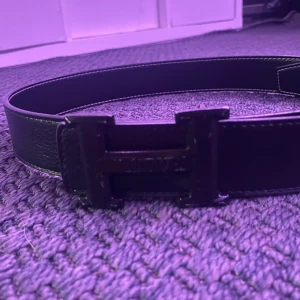 Svart skärp med H-spänne i skinn - Stilrent svart bälte i skinn med ett brett H-format spänne framtill. Bältet har en klassisk design med synliga vita sömmar längs kanterna och en slät, lätt texturerad yta. Perfekt accessoar för att lyfta din outfit med en clean och modern look.