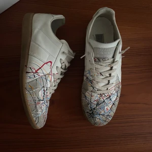 Maison Margiela sneakers med färgstänk - Maison Margiela sneakers i vitt skinn med unikt färgstänkt mönster i blått, rött, gult och svart. Skorna är storlek 43, och allt orginalt med följer t.ex som box och dustbags, de ända jag inte har är kvitto tyvärr, men de är äkta och de kan ja garantera, priset är inte hugget i sten så kom med pris för salg, OBS tar även byten mot stor i storleken 40-41!!!