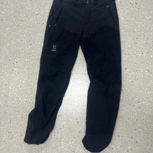 Svarta täckbyxor från Haglöfs - Svarta täckbyxor från Haglöfs med dragkedjeförsedda fickor och förstärkta knän. Perfekta för utomhusaktiviteter och har en sportig look. Byxorna har normal passform och är tillverkade i slitstarkt syntetmaterial.  Märket gabbro
