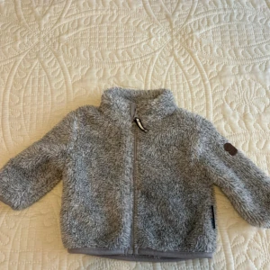 Grå teddyjacka från On The Peak - Mysig grå teddyjacka från On The Peak i storlek 62. Jackan har fluffig yta, hög krage och dragkedja framtill. Perfekt för kyliga dagar och har långärmade ärmar. Snygg detalj med logopatch på ena ärmen.