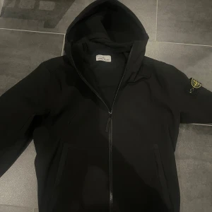 Svart vindjacka från Stone Island - Svart vindjacka från Stone Island med huva och dragkedja framtill. Jackan har ikonisk patch på ärmen och är tillverkad i teknisk polyester med soft shell-R e.dye® teknologi som gör den både vindtät och vattenavvisande. Snygg och funktionell för dig som gillar streetwear.