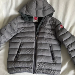 Colmar grå pufferjacka  - Säljer en grå pufferjacka från Colmar med huva. Perfekt till höst, vinter och vår. Storlek 52, jag är 189 och den sitter lite kort på mig. Skulle säga att den sitter som en M.