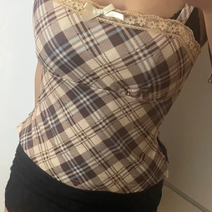 Rutig korsett-topp med spets och rosett - Supersöt korsett-topp med brunt och beige rutigt mönster, dekorerad med spetskant och en liten rosett framtill. Axelbandsmodell med figurnära passform, perfekt för dig som gillar vintagevibes och detaljer. Materialet känns mjukt och har en lätt stretch.