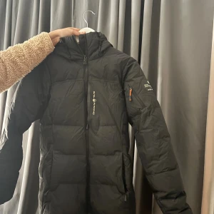 Svart pufferjacka Sail Racing - Svart pufferjacka från Sail Racing med huva och hel dragkedja framtill. Jackan har en ficka med dragkedja på ärmen och logga på bröstet. Perfekt för kalla dagar och har en clean, sportig look.