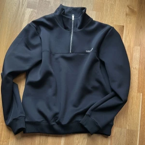Svart half zip tröja Les Deux - Svart långärmad tröja från Les Deux med hög krage och half zip-dragkedja framtill. Tröjan har en stilren look med diskret logga på bröstet och ribbade muddar vid ärmslut och nederkant. Perfekt för lager-på-lager och chill vardagsstil.