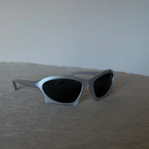 Grå/Silvriga solglasögon med svart glas - Snygga solglasögon med silvrigt, plastigt cat-eye båge och svarta linser. Den futuristiska formen ger en cool vibe och passar dig som vill sticka ut. Perfekt accessoar för att lyfta din stil med en modern touch. Aldrig använda.