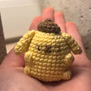 Virkad pompompurin sanrio - Denna produkt är en handvirkad figur, troligen inspirerad av en karaktär från Sanrio,  Pompompurin. Den är gjord av gult och brunt garn och är liten nog att rymmas i handen.  Perfekt som dekoration, present eller att ha som nyckelkedja. 