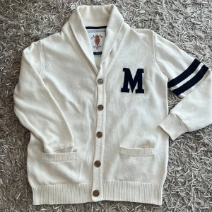 Vit kofta från Morris med marinblå detaljer - Snygg vit kofta från Morris med marinblå M på bröstet och två marinblå ränder på ena ärmen. Koftan har knappar framtill, två fickor och en klassisk krage. Perfekt för dig som gillar preppy stil och vill sticka ut lite extra. Pris kan diskuteras!