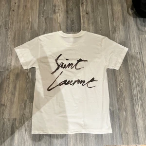  t-shirt med tryck - Säljer en vit t-shirt från Saint Laurent med stort handskrivet logotryck i svart på ryggen. T-shirten har klassisk rund hals och korta ärmar. Materialet är mjuk bomull som känns skönt mot huden. Perfekt för dig som gillar stilrena designer med en lyxig touch.