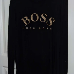 Svart långärmad tröja Hugo Boss - Svart långärmad tröja från Hugo Boss med stort guldigt BOSS-tryck på bröstet. Enkel och stilren design som passar perfekt till jeans eller dressade byxor. Mjuk och skön bomullskvalitet.