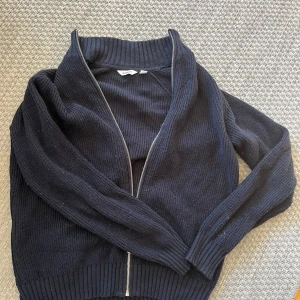 Mörkblå stickad kofta från Dobber - Säljer en svart stickad kofta från Dobber med grov ribbad krage och muddar. Koftan har en snygg silverfärgad dragkedja framtill och långärmade ärmar. Perfekt för lager-på-lager och chill stil. Passar dig som gillar klassiska och enkla plagg. Har en del ludd på sig, men ska enkelt gå att få bort