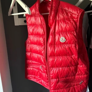 Röd dunväst från Moncler - Snygg röd dunväst från Moncler med quiltad design och klassisk logotyp på bröstet. Västen har dragkedja framtill och två praktiska fickor med dragkedja. Perfekt att slänga över en hoodie för en cool look. Materialet är glansigt och lätt, vilket gör den enkel att bära. Allt fås med och prisförslag uppskattas!!