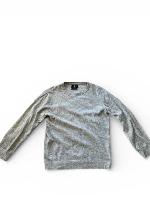 Grå stickad tröja från SC - Grå stickad tröja från SC med rund hals och broderad logga på bröstet. Tröjan har långärmade ärmar och ribbade muddar vid ärmslut och nederkant. Perfekt för kyliga dagar och enkel att matcha med jeans eller chinos. Tröjan har några små hål vid armslutet men det syns inte vid användning. Måtten på tröjan är 70cm Längd och 53cm Bredd.